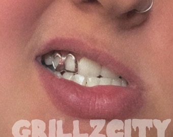 May include: A silver-coloured metal tooth grillz piece covering two teeth, positioned on the upper right side of the mouth. The image features the text GRILLZCITY in a white, dripping-style font overlaid at the bottom of the frame.