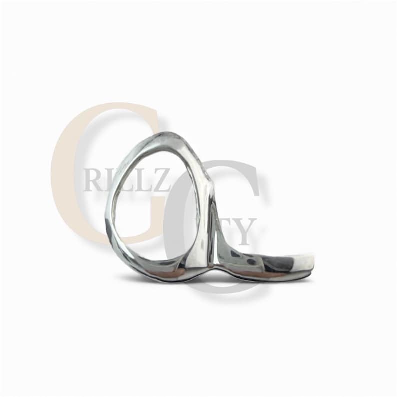 May include: A silver-toned ring with an abstract design. The ring features a rounded oval shape connected to a flat, curved band. The surface of the ring appears smooth and reflective, with a polished finish. The ring is set against a white background.