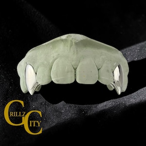 May include: Silver-colored fang grillz on a green dental mold. The grillz are highly polished and reflect light. The logo "GRILLZ CITY" is in gold lettering at the bottom left corner against a black background.