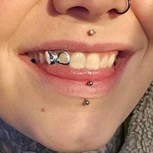 May include: Close-up of a person's mouth with a silver tooth cap and multiple piercings. The silver tooth cap is on the upper left tooth. The piercings are on the lip and nose.