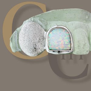 May include: A silver-toned dental grill with an opal stone set in a rectangular shape. The grill is placed on a light green dental mold, with a silver textured ball on the left side. The background is a muted brown with a gold-colored letter "C".