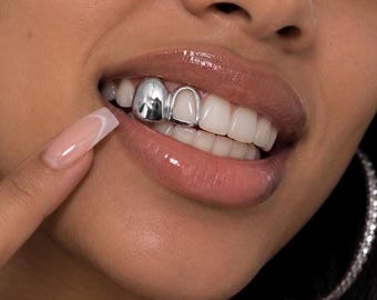 May include: A silver-coloured tooth grill covering two upper teeth, featuring one solid polished cap and one open-frame cap. The metallic accessory is positioned on the upper teeth, showcasing a reflective surface and a smooth, rounded design.