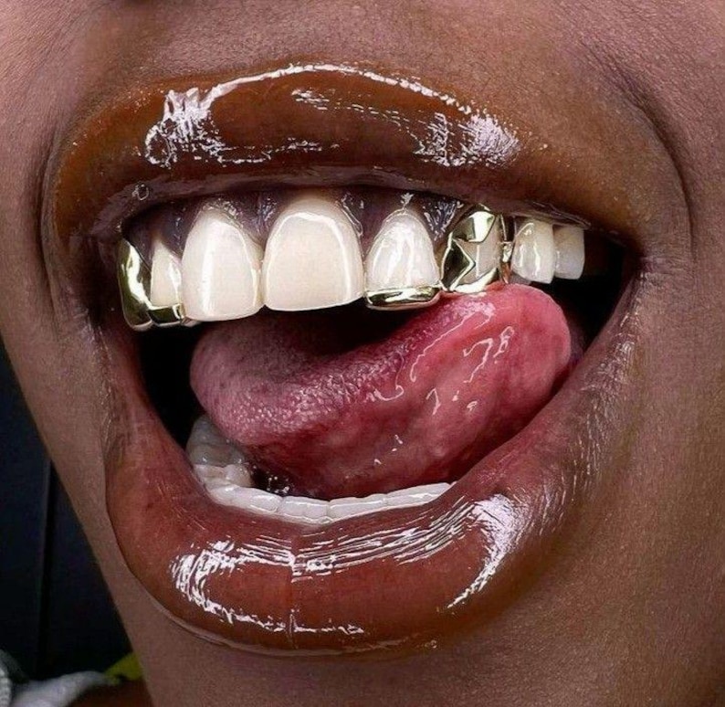 May include: Close-up of a mouth with glossy, dark brown lips and white teeth. Gold grillz with star designs are visible on the upper teeth. The tongue is extended, showing a pinkish-red color.