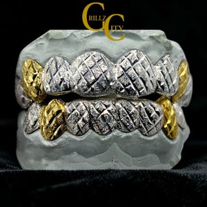 May include: Silver and gold-toned grillz with a diamond-cut pattern. The grillz are displayed on a grey mold, with the text "RILLZ CITY" in gold at the top. The grillz have a shiny, reflective surface.