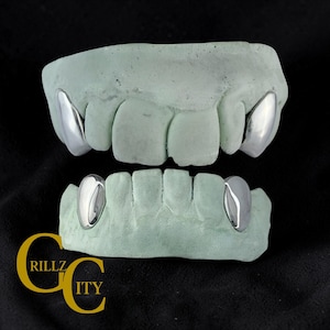 May include: A set of silver-colored fang grillz, displayed on light green dental molds. The grillz are highly polished and reflect light. The logo "RILLZ CITY" is visible in the bottom left corner.