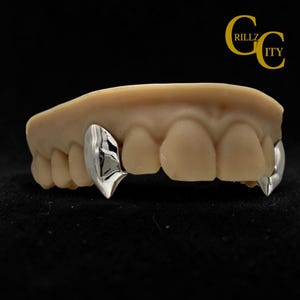 May include: A dental model of upper teeth featuring two polished silver-colored fang caps placed over the canine teeth. The model is beige and shows a full row of teeth. The text GRILLZ CITY is visible in the upper right corner in yellow.
