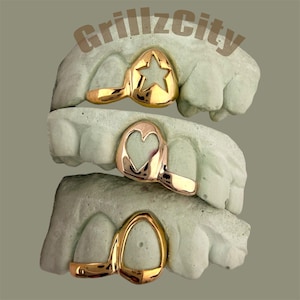 May include: Gold-colored grillz with star, heart, and oval designs are displayed on dental molds. The text "GrillzCity" is visible at the top. The grillz are shown in a stacked arrangement, showcasing the variety of styles.