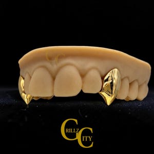 May include: A dental model of upper teeth featuring two polished gold canine caps. The gold caps are pointed and reflective. The text GRILLZ CITY is visible in gold lettering at the bottom and faintly on the gum line of the model.