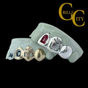 May include: Silver and gold-toned grillz with cross designs, displayed on dental molds. The grillz feature a mix of gold and silver finishes, with a prominent cross motif. The image includes the text "GRILLZ CITY" in gold.