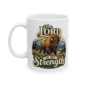 May include: A white ceramic mug featuring a graphic of a brown bison standing in a mountain landscape. The text reads The LORD IS MY Strength and Psalm 28:7. The design includes pine trees, rocky terrain, and a sunburst background in earth tones.