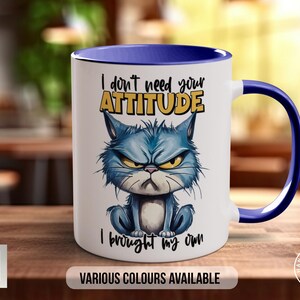 May include: White ceramic mug with a blue rim and handle. The mug features a cartoon cat with a grumpy expression and the text "I don't need your ATTITUDE I brought my own".