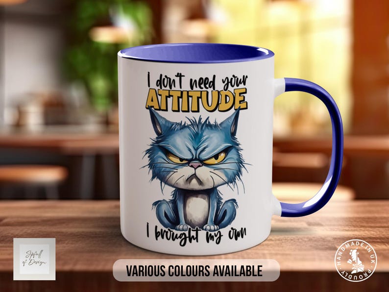 May include: White ceramic mug with a blue rim and handle. The mug features a cartoon cat with a grumpy expression and the text "I don't need your ATTITUDE I brought my own".