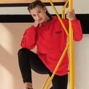 Può includere: Una giacca rossa con cappuccio, chiusura a zip e il testo "Don't Trust, Verify" ricamato sul davanti. La giacca è indossata da una persona in piedi su un ponteggio giallo.
