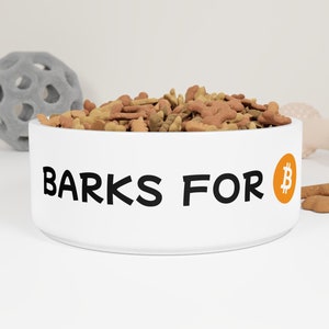May include: White ceramic dog bowl filled with dog food. The bowl has the text "BARKS FOR" in black, with an orange Bitcoin symbol. A gray ball and a light pink bone-shaped toy are in the background.