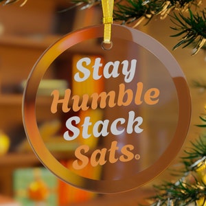 May include: A clear, round Christmas ornament with the words "Stay Humble Stack Sats" in white and orange lettering. The ornament has a gold ribbon for hanging and is surrounded by a brown border. The background is blurred.