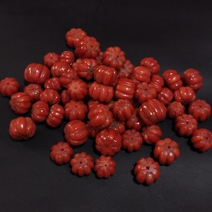 May include: A pile of small, carved red pumpkin beads. The beads are of varying sizes and feature detailed ridges and a hole for threading. These decorative beads are suitable for jewellery making and craft projects.