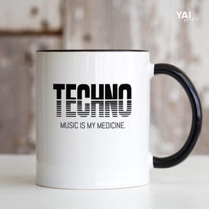 Könnte beinhalten: Ein weißer Keramikbecher mit schwarzem Henkel und Rand. Auf der Vorderseite steht das Wort TECHNO in einer fetten, schwarzen, horizontal gestreiften Schrift, darunter der Text MUSIC IS MY MEDICINE in einer einfachen, klaren, schwarzen serifenlosen Schrift.