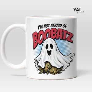 Puede incluir: Una taza de cerámica blanca con un gráfico de un fantasma blanco flotando sobre un grupo de cogollos de cannabis verdes y naranjas. El texto sobre el fantasma dice I'M NOT AFRAID OF BOOBATZ en letras negras y rojas sobre un fondo azul punteado.