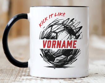 Könnte beinhalten: Ein weißer Keramikbecher mit schwarzem Henkel und schwarzem Innenraum. Auf der Vorderseite befindet sich eine schwarze Grafik eines Fußballs mit Bewegungslinien, der rote Schriftzug KICK IT LIKE und ein rotes Banner mit VORNAME.