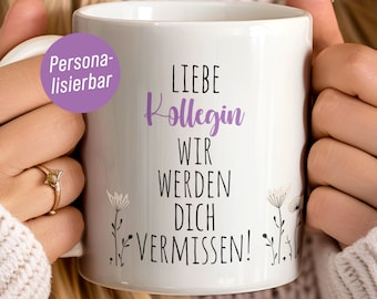 Puede incluir: Taza de cerámica blanca con una inscripción morada que dice "Liebe Kollegin Wir Werden Dich Vermissen!" con un diseño floral. La taza está siendo sostenida por una mano.