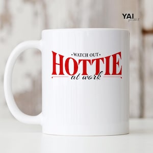 Op de afbeelding: Een witte keramische mok met de rode tekst "WATCH OUT" boven het grote rode woord "HOTTIE" en de zwarte cursieve tekst "at work" eronder. De mok heeft een standaard handvat aan de linkerkant en een gladde, glanzende afwerking over het geheel.