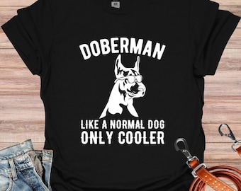 Op de afbeelding: Zwart T-shirt met witte tekst die "Doberman Like a Normal Dog Only Cooler" luidt en een wit silhouet van een Dobermann met een zonnebril.