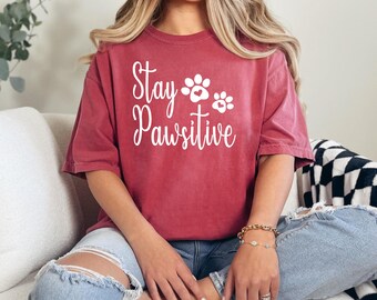 Peut inclure: Une femme portant un t-shirt rouge foncé avec le texte "Stay Pawsitive" et deux empreintes de pattes avec des cœurs. Elle est assise sur un canapé blanc avec une couverture à carreaux noir et blanc.