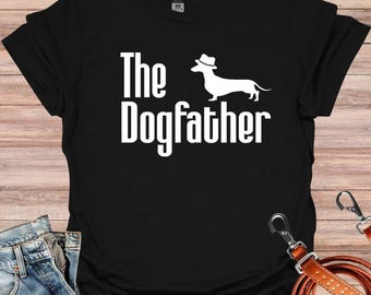 May include: Black t-shirt with white text that reads "The Dogfather" and a white silhouette of a dachshund wearing a fedora.
