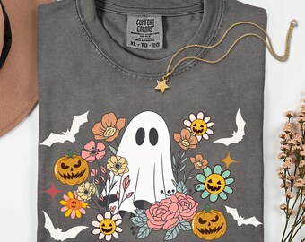 Op de afbeelding: Een grijs Comfort Colors XL T-shirt met een Halloween-ontwerp. Het ontwerp toont een witte geest, pompoenen, bloemen en vleermuizen. Een gouden sterrenketting is over het shirt gedrapeerd.