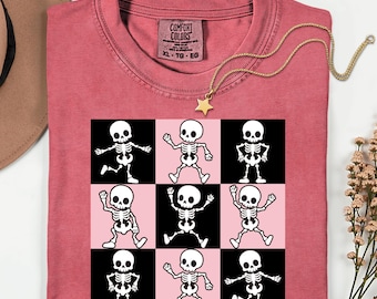 Puede incluir: Una camiseta de color sandía con cuello redondo. La camiseta presenta una cuadrícula de nueve ilustraciones de esqueletos bailando en blanco y negro sobre cuadrados rosas y negros. Un collar con colgante de estrella dorada está sobre la camiseta.