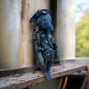 Puede incluir: Un juguete de peluche azul y gris con un pico largo, que lleva un gorro de aviador de cuero y gafas, y adornado con cadenas y engranajes de metal. El juguete está de pie en un estante de madera.