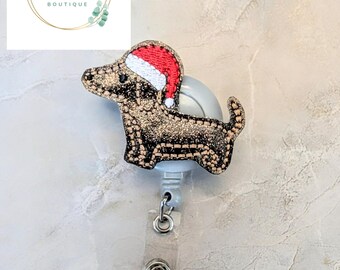 May include: A glittery dachshund badge reel with a Santa hat. The dog is black and gold with a red and white hat. The reel has a clear plastic strap and a metal clip. The logo for Ivy Nicole Boutique is in the upper left corner.