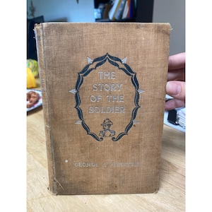 May include: A vintage book titled "The Story of the Soldier" with a tan cover. The title is in silver lettering within a decorative blue and silver frame. The author's name, George A. Birmingham, is printed at the bottom.