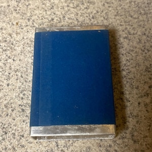 Puede incluir: Una caja rectangular cubierta de terciopelo azul con ribete de metal plateado. La caja est&aacute; cerrada y parece ser un estuche o soporte peque&ntilde;o. El terciopelo es de un color azul intenso y rico.
