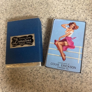 Puede incluir: Juego de cartas Duratone vintage en un estuche azul con ribete plateado. El estuche tiene el texto "Plastic Coated Playing Cards". La baraja abierta presenta una ilustraci&oacute;n pin-up con el texto "Compliments of Couie Erickson Standard Oil".