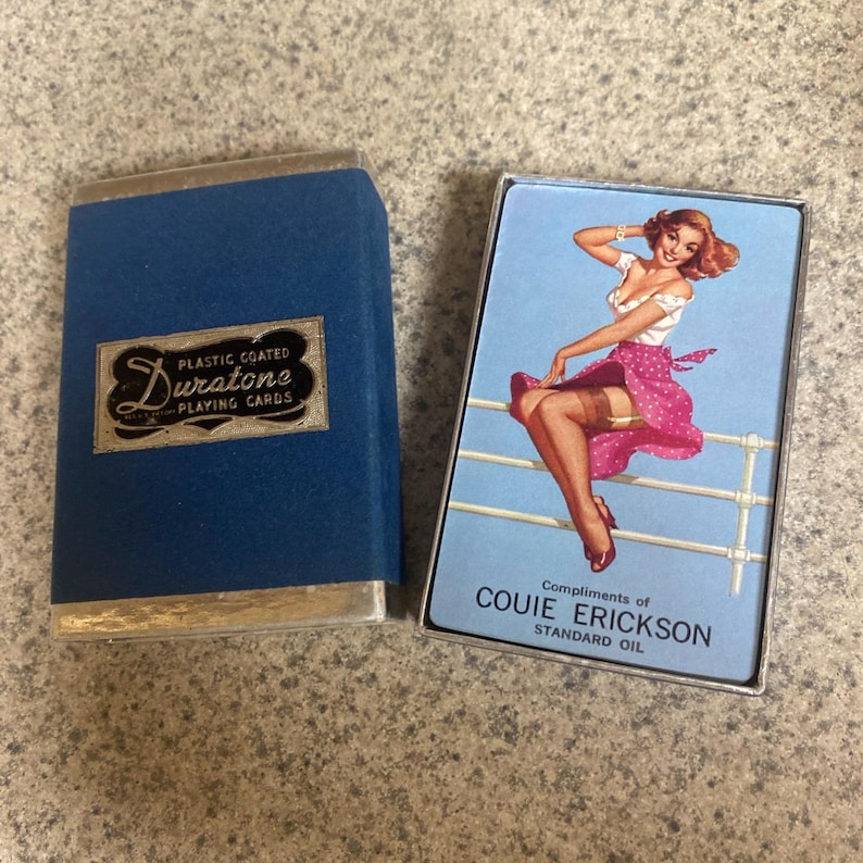 Puede incluir: Juego de cartas Duratone vintage en un estuche azul con ribete plateado. El estuche tiene el texto "Plastic Coated Playing Cards". La baraja abierta presenta una ilustraci&oacute;n pin-up con el texto "Compliments of Couie Erickson Standard Oil".