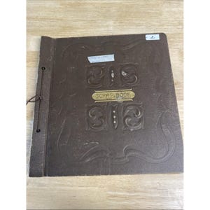 Puede incluir: Un álbum de recortes marrón vintage con diseños florales en relieve y las palabras "SCRAP BOOK" en una placa dorada. El álbum está encuadernado con un cordón marrón y tiene una pequeña etiqueta blanca con texto.