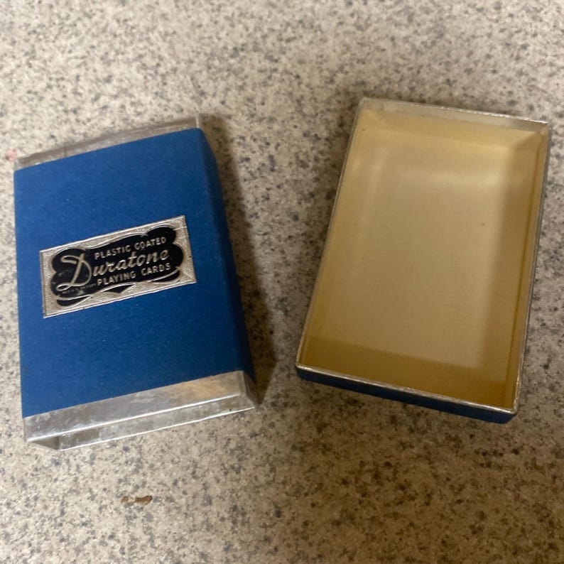 Puede incluir: Una caja azul de naipes Duratone con ribete plateado, abierta junto a su interior beige vac&iacute;o. La caja tiene las palabras "PLASTIC COATED Duratone PLAYING CARDS" en un emblema plateado.