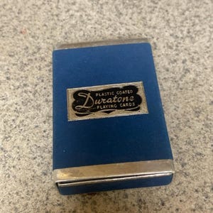 Puede incluir: Un estuche azul para naipes Duratone con bordes plateados. El estuche tiene forma rectangular y presenta una etiqueta plateada con las palabras "Duratone" y "Plastic Coated Playing Cards". El estuche est&aacute; cerrado.