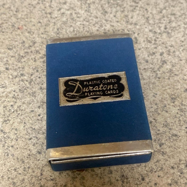 Puede incluir: Un estuche azul para naipes Duratone con bordes plateados. El estuche tiene forma rectangular y presenta una etiqueta plateada con las palabras "Duratone" y "Plastic Coated Playing Cards". El estuche est&aacute; cerrado.