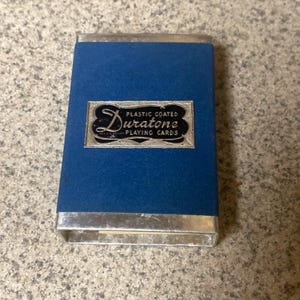 Puede incluir: Un estuche de naipes vintage con exterior de terciopelo azul y ribete met&aacute;lico plateado. El estuche presenta una etiqueta rectangular con el texto "Duratone Plastic Coated Playing Cards" en una fuente decorativa.