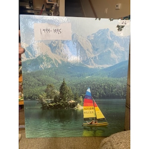 May include: A scenic landscape print featuring a sailboat on a lake with a mountain backdrop. The sailboat has a colorful sail. The text "1994-1995" is visible on the top left. A small island with trees is in the center of the lake.