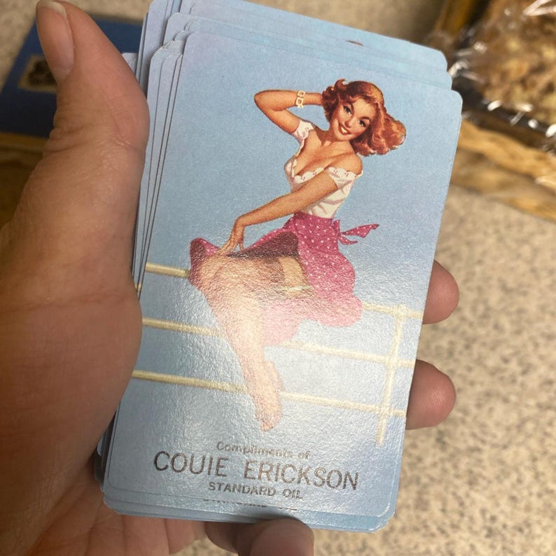 Puede incluir: Un mazo de cartas de juego azul claro con una ilustraci&oacute;n vintage de una pin-up. La mujer lleva un top blanco y una falda rosa con lunares. El texto "Compliments of Couie Erickson Standard Oil" est&aacute; impreso en la parte inferior.