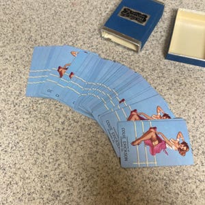 Puede incluir: Una baraja de cartas de juego azul claro con arte pin-up, abanicada sobre una superficie moteada. Las cartas est&aacute;n etiquetadas como "Couie Erickson" y van acompa&ntilde;adas de una caja azul con ribetes plateados.
