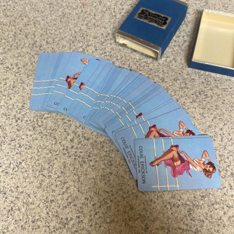Puede incluir: Una baraja de cartas de juego azul claro con arte pin-up, abanicada sobre una superficie moteada. Las cartas est&aacute;n etiquetadas como "Couie Erickson" y van acompa&ntilde;adas de una caja azul con ribetes plateados.