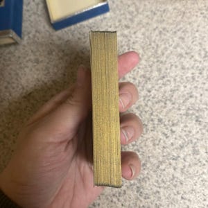Puede incluir: Un mazo de cartas con bordes dorados se sostiene en una mano. Las cartas son rectangulares y est&aacute;n apiladas. En el fondo se ve una caja azul con el interior dorado.