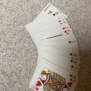 Puede incluir: Un mazo de naipes abanicado, mostrando varios palos y valores. La Reina de Corazones es visible en la parte inferior, con otras cartas que muestran corazones, picas y tr&eacute;boles. La carta del Joker tambi&eacute;n es visible. Las cartas est&aacute;n sobre una superficie gris claro.