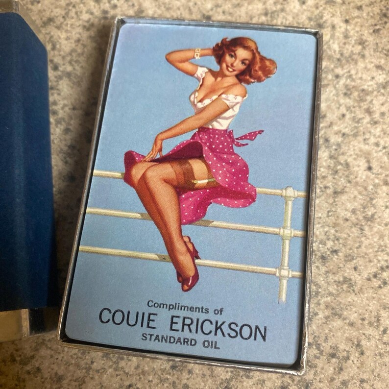Puede incluir: Una carta de juego vintage con una ilustraci&oacute;n pin-up de una mujer con top blanco y falda de lunares rosa, apoyada en una barandilla blanca. La tarjeta tiene el texto "Compliments of COUIE ERICKSON STANDARD OIL" sobre un fondo azul claro.