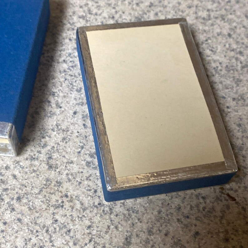 Puede incluir: Una caja rectangular azul con borde plateado e interior crema. La caja est&aacute; cerrada, con un borde plateado visible. Otra caja azul es parcialmente visible en la imagen.