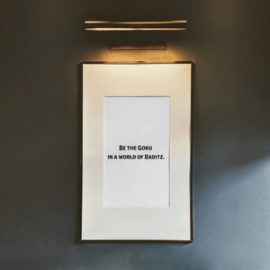 May include: A framed print with the text "Be the Goku in a world of Raditz." The print is hanging on a dark gray wall with a light fixture above it.
